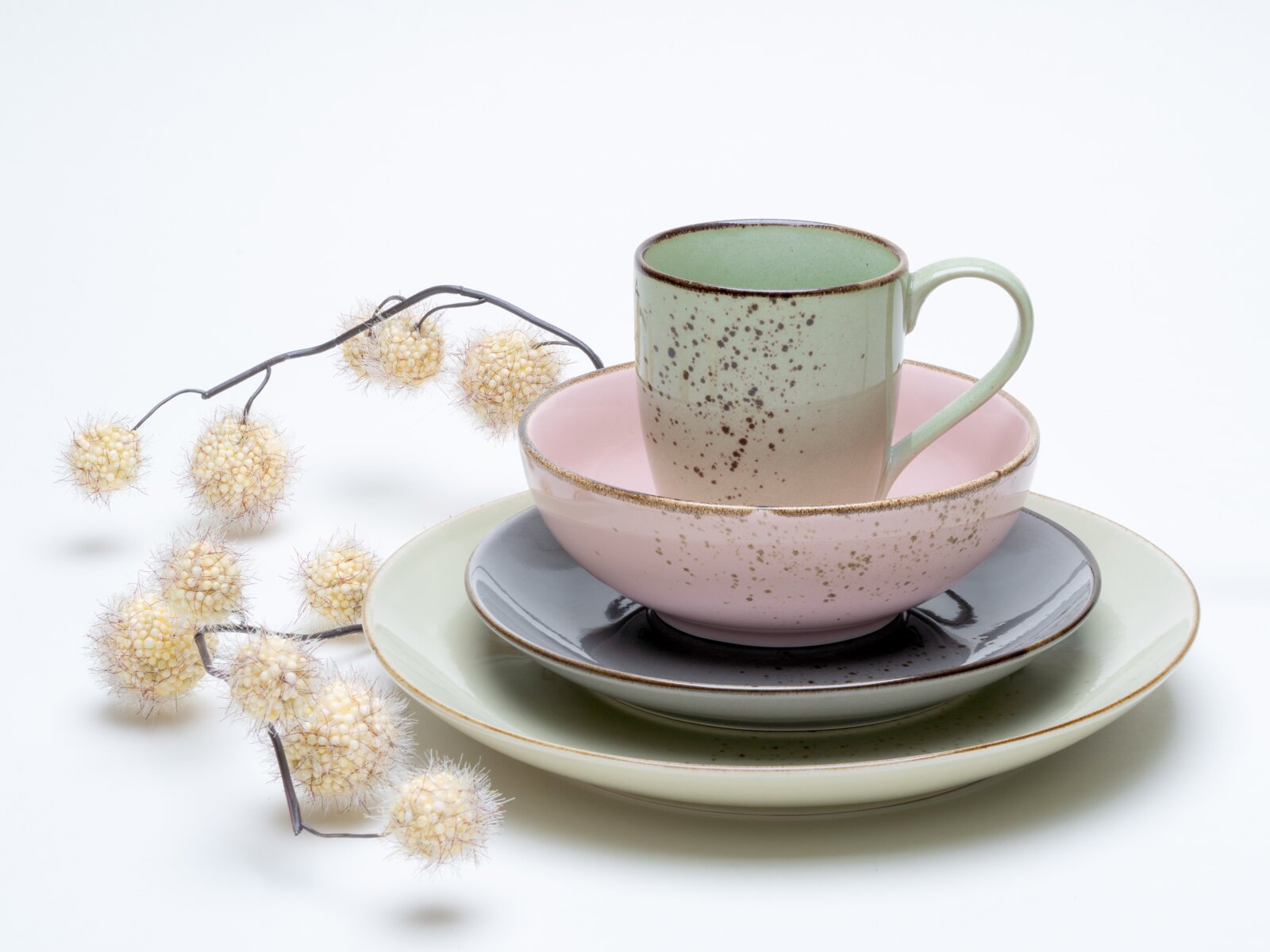 Pastellfarbenes, gesprenkeltes Geschirr-Set mit Tasse, Schale und Tellern auf weißem Hintergrund, dekoriert mit Trockenblumen, fotografiert in leichter Seitenansicht (3/4-Perspektive) von vorne.
