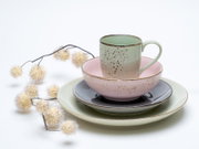 Pastellfarbenes, gesprenkeltes Geschirr-Set mit Tasse, Schale und Tellern auf weißem Hintergrund, dekoriert mit Trockenblumen, fotografiert in leichter Seitenansicht (3/4-Perspektive) von vorne.