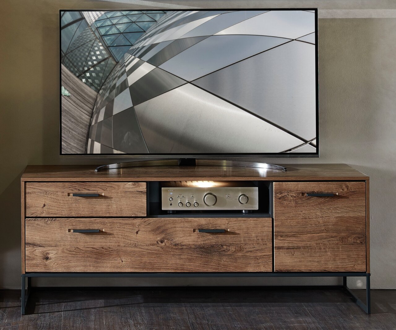 TV-Lowboard MANHATTAN aus Holz mit Metallakzenten, frontal fotografiert, mit Schubladen und Stauraum, darauf ein moderner Flachbildfernseher.