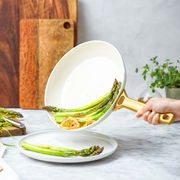 Nahaufnahme einer weißen Bratpfanne mit goldfarbenem Griff, in der Spargel und Zitronenscheiben serviert werden. Die Pfanne wird über einem weißen Teller gehalten.