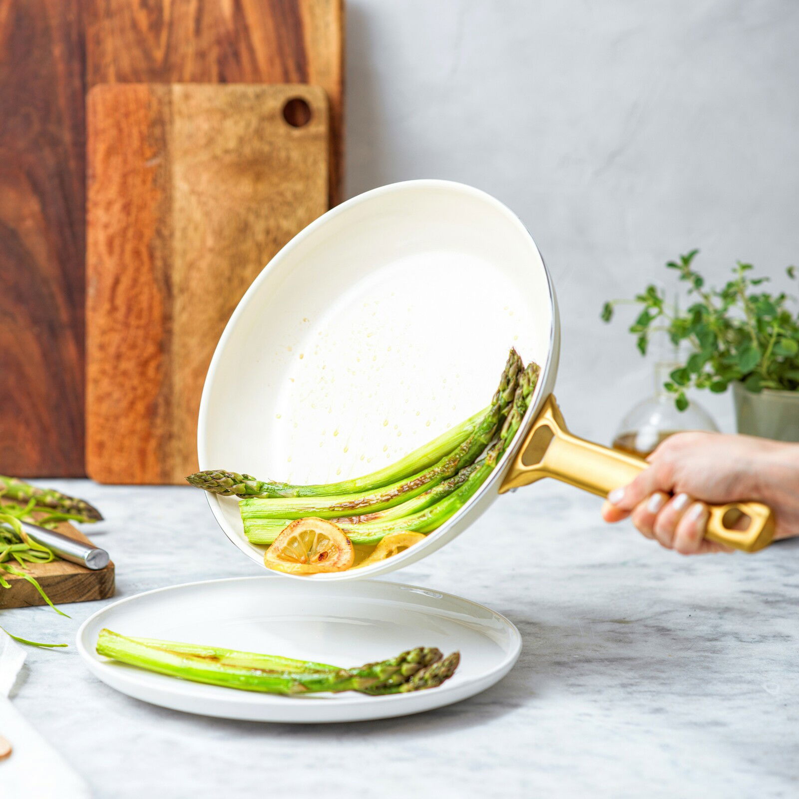 Nahaufnahme einer weißen Bratpfanne mit goldfarbenem Griff, in der Spargel und Zitronenscheiben serviert werden. Die Pfanne wird über einem weißen Teller gehalten.