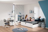 Zwei junge Frauen entspannen auf einer Universalliege in einem modernen, hellen Zimmer mit Schreibtisch und Regalen, seitliche Perspektive.