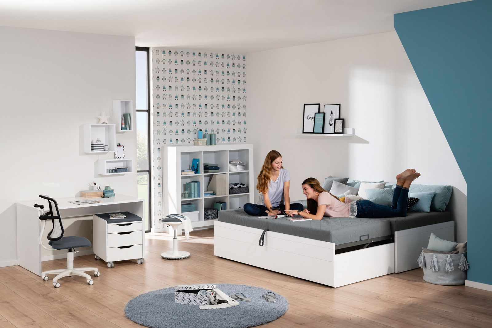 Zwei junge Frauen entspannen auf einer Universalliege in einem modernen, hellen Zimmer mit Schreibtisch und Regalen, seitliche Perspektive.