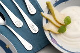 Kinderbesteck-Set mit vier Teilen, bestehend aus Löffel, Gabel und zwei Messern, verziert mit kindgerechten Motiven wie einer Katze und Figuren. Das Besteck liegt auf einem blauen Tuch neben einer Schale mit Joghurt und Birnenscheiben. Perspektive von obe
