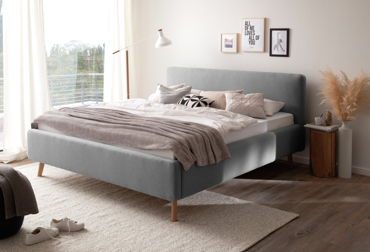 Modernes Polsterbett in Grau mit Holzfüßen, seitliche Perspektive, dekoriert mit Kissen und Decke in einem hellen Schlafzimmer.