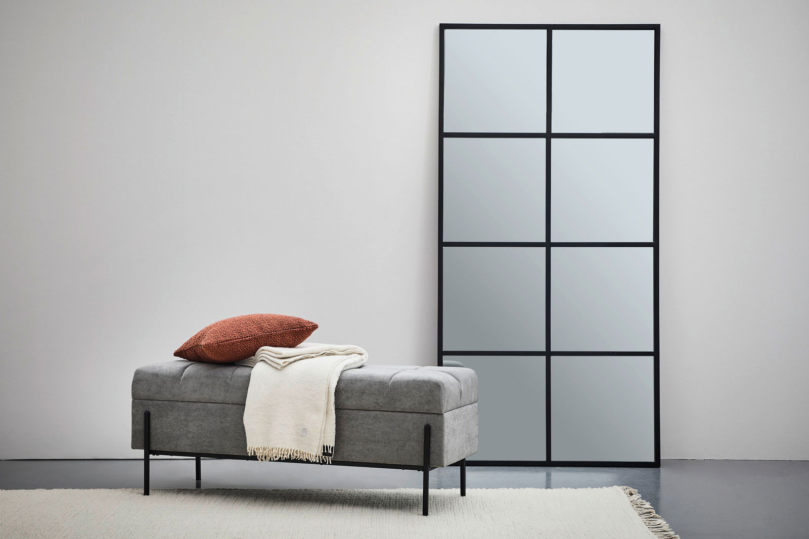 Graue Hockerbank mit Stauraum, darauf ein rotes Kissen und eine cremefarbene Decke, seitliche Perspektive.