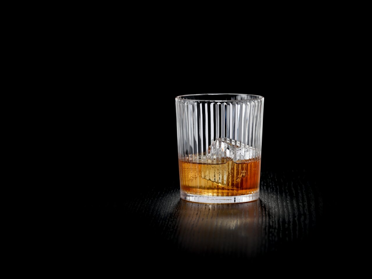 Nachtmann Becher-Set 2-tlg. APERITIVO Ein elegantes, geripptes Glas mit einem Getränk und Eiswürfeln, auf einem dunklen Tisch aus der Seitenperspektive fotografiert.