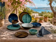 Serviescollectie met geglazuurde borden, vakborden en kommen in blauw, turquoise, bruin en crème, vanuit schuin vooraanzicht gepresenteerd op een houten tuintafel aan zee