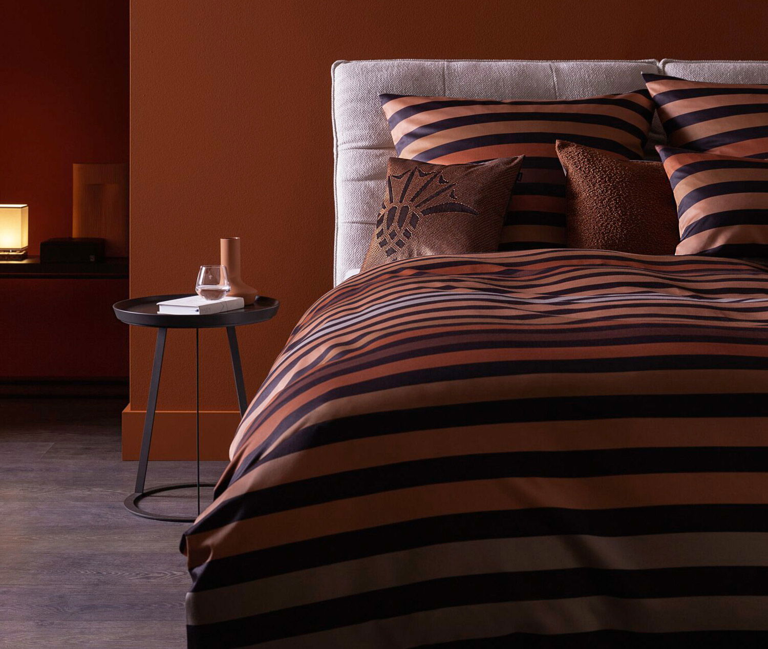 Gemütliches Bett mit gestreifter brauner und schwarzer Bettwäsche, akzentuiert durch dekorative Kissen, neben einem kleinen Tisch mit einem Glas und einem Buch.