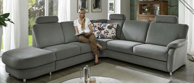 Beldomo System Ecksofa TAULON-S Graues Ecksofa TAULON-S in einem modernen Wohnzimmer, mit einer Person, die darauf sitzt. Perspektive von vorne links.
