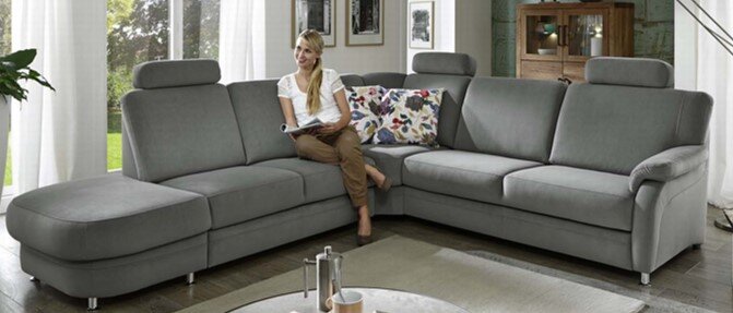 Beldomo System Ecksofa TAULON-S Graues Ecksofa TAULON-S in einem modernen Wohnzimmer, mit einer Person, die darauf sitzt. Perspektive von vorne links.