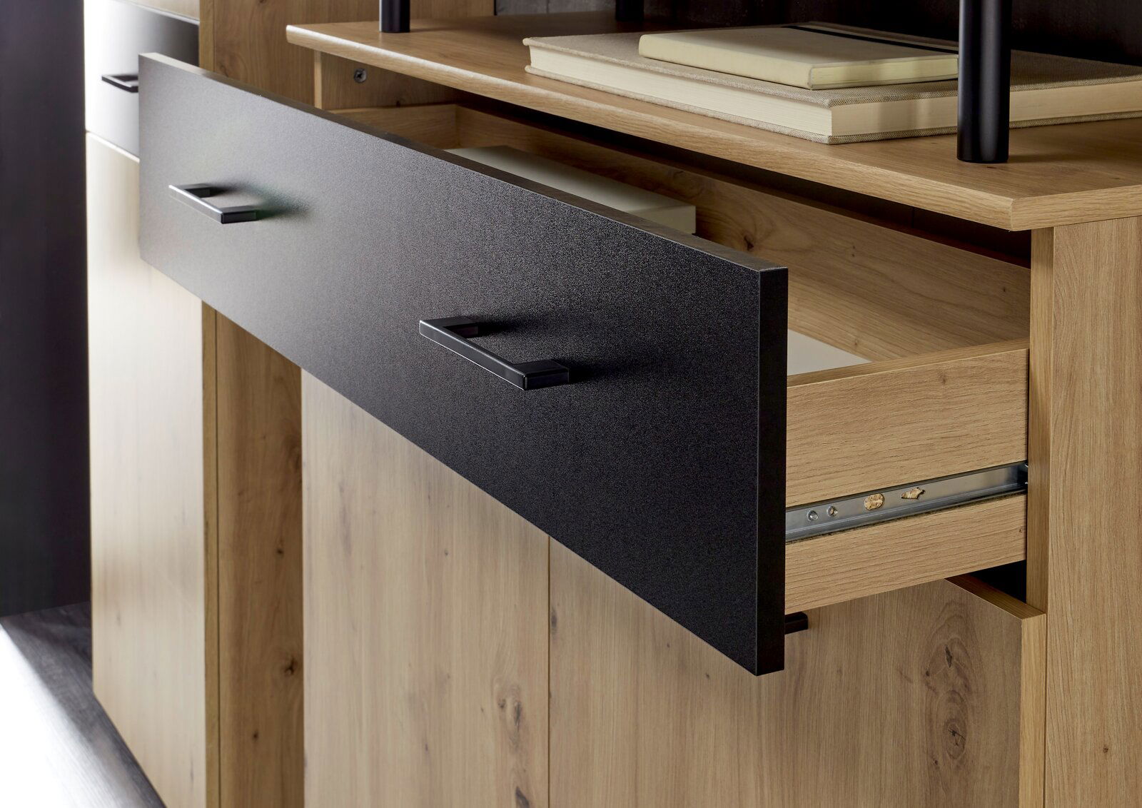 Nahaufnahme einer geöffneten Schublade eines Garderoben-Sets aus Holz mit schwarzer Front und modernen Griffen, seitliche Perspektive.