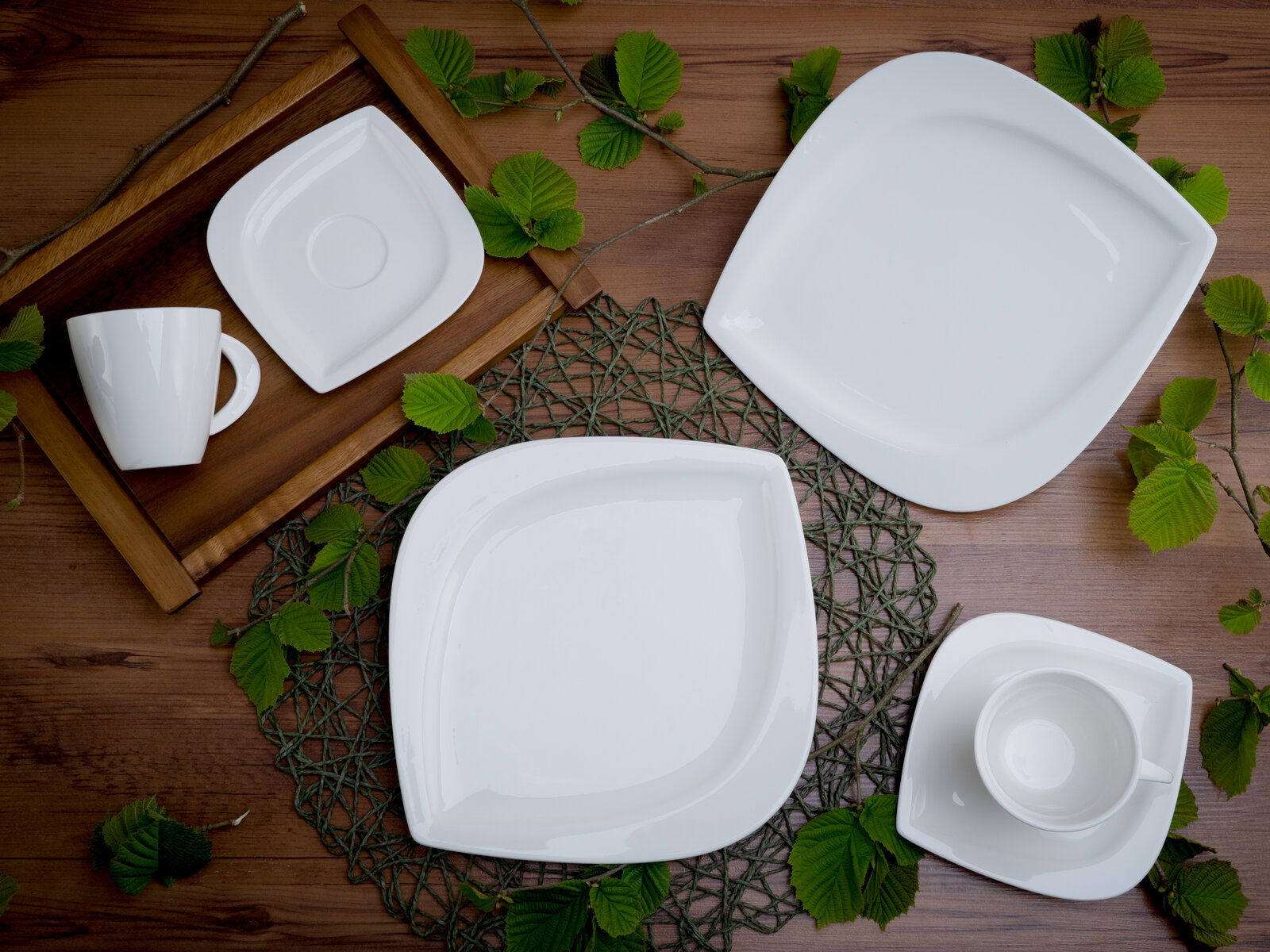 Vogelperspektive auf ein elegantes, weißes Kaffeeservice-Set mit Tassen, Untertassen und Tellern, dekoriert mit grünen Blättern auf einem Holztisch.
