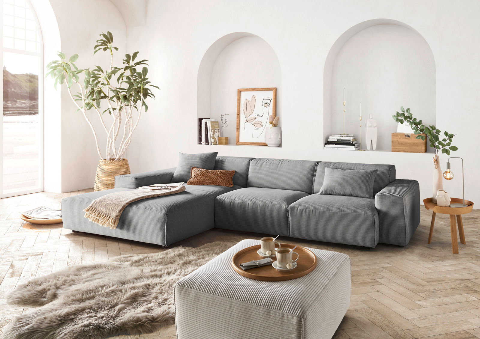 Graue Rundecke mit Ottomane in einem modernen Wohnzimmer, seitliche Perspektive. Dekoriert mit Kissen, Decke und Pflanzen.