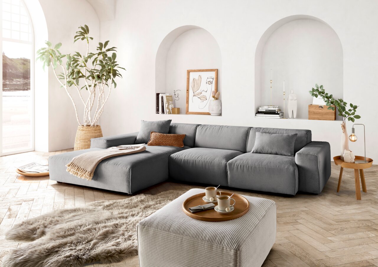 Graue Rundecke mit Ottomane in einem modernen Wohnzimmer, seitliche Perspektive. Dekoriert mit Kissen, Decke und Pflanzen.