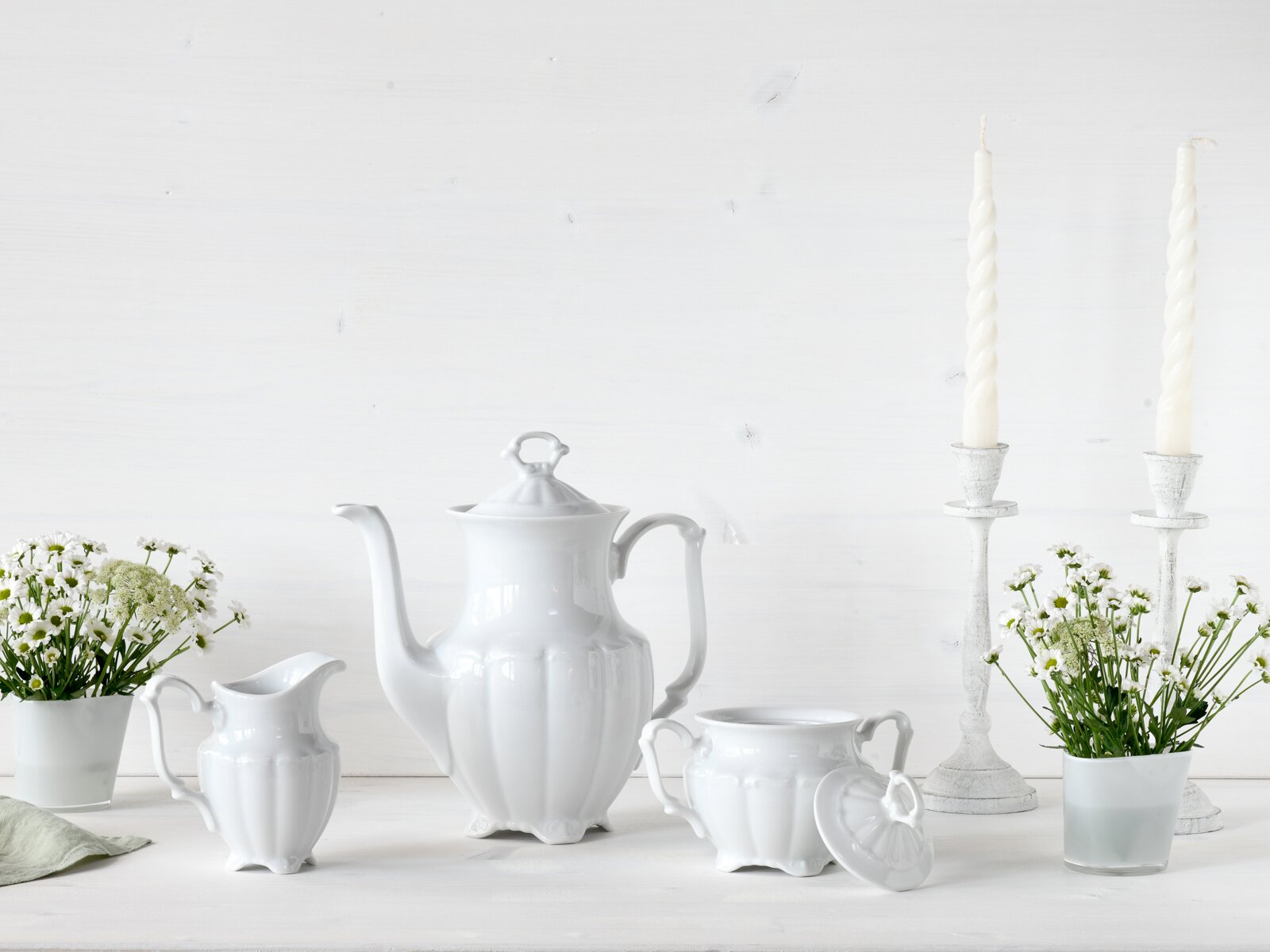 Weiße Porzellan-Kaffeekanne mit passendem Milchkännchen und Zuckerdose mit Deckel auf hellem Tisch, dekoriert mit Blumen und Kerzenhaltern, Frontansicht auf Augenhöhe.