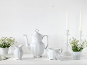 Weiße Porzellan-Kaffeekanne mit passendem Milchkännchen und Zuckerdose mit Deckel auf hellem Tisch, dekoriert mit Blumen und Kerzenhaltern, Frontansicht auf Augenhöhe.