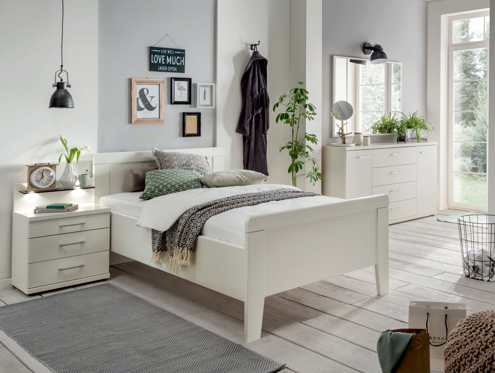Venda Nachtkastje BERN Moderne witte nachtkastje met drie lades, naast een bed in een stijlvol ingerichte slaapkamer, gezien vanaf de zijkant.