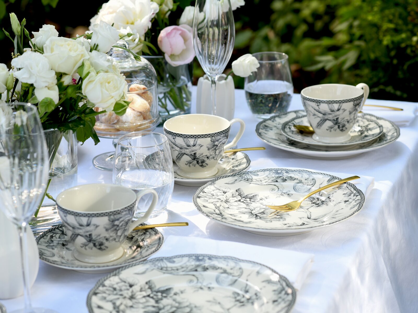 Elegantes Kaffee- und Tafelservice aus weißem Porzellan mit schwarz-grauem floralen Muster auf gedecktem Tisch mit goldfarbenem Besteck, Wassergläsern und Blumenarrangement im Garten, Perspektive: leicht schräg von oben (3/4-Ansicht)