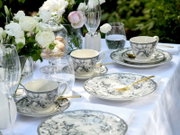 Elegantes Kaffee- und Tafelservice aus weißem Porzellan mit schwarz-grauem floralen Muster auf gedecktem Tisch mit goldfarbenem Besteck, Wassergläsern und Blumenarrangement im Garten, Perspektive: leicht schräg von oben (3/4-Ansicht)
