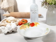 Weiße Pastaschale mit Tomaten- und Pasta-Motiv am Rand auf hellem Küchentisch, aufgenommen in leicht erhöhter Vorderansicht; im Hintergrund Nudelholz, Olivenölflasche, Eier und Tomaten.