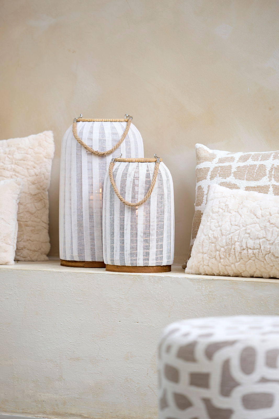 Zwei Windlichter mit weißen Stoffstreifen und Seilgriffen, auf einem hellen Podest platziert, umgeben von dekorativen Kissen. Seitliche Perspektive.
