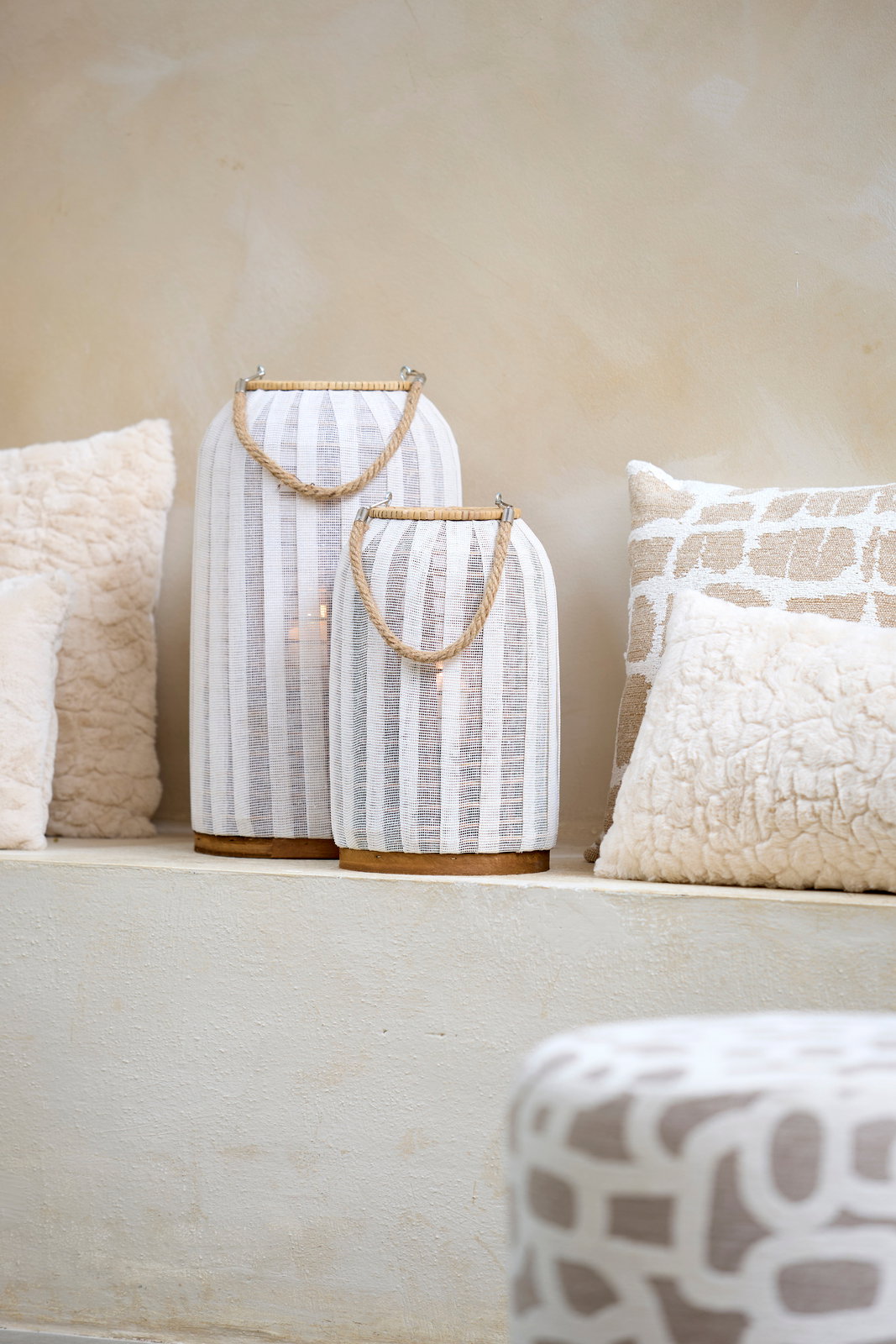 Light&Living Windlicht YLINA 35cm Zwei Windlichter mit weißen Stoffstreifen und Seilgriffen, auf einem hellen Podest platziert, umgeben von dekorativen Kissen. Seitliche Perspektive.