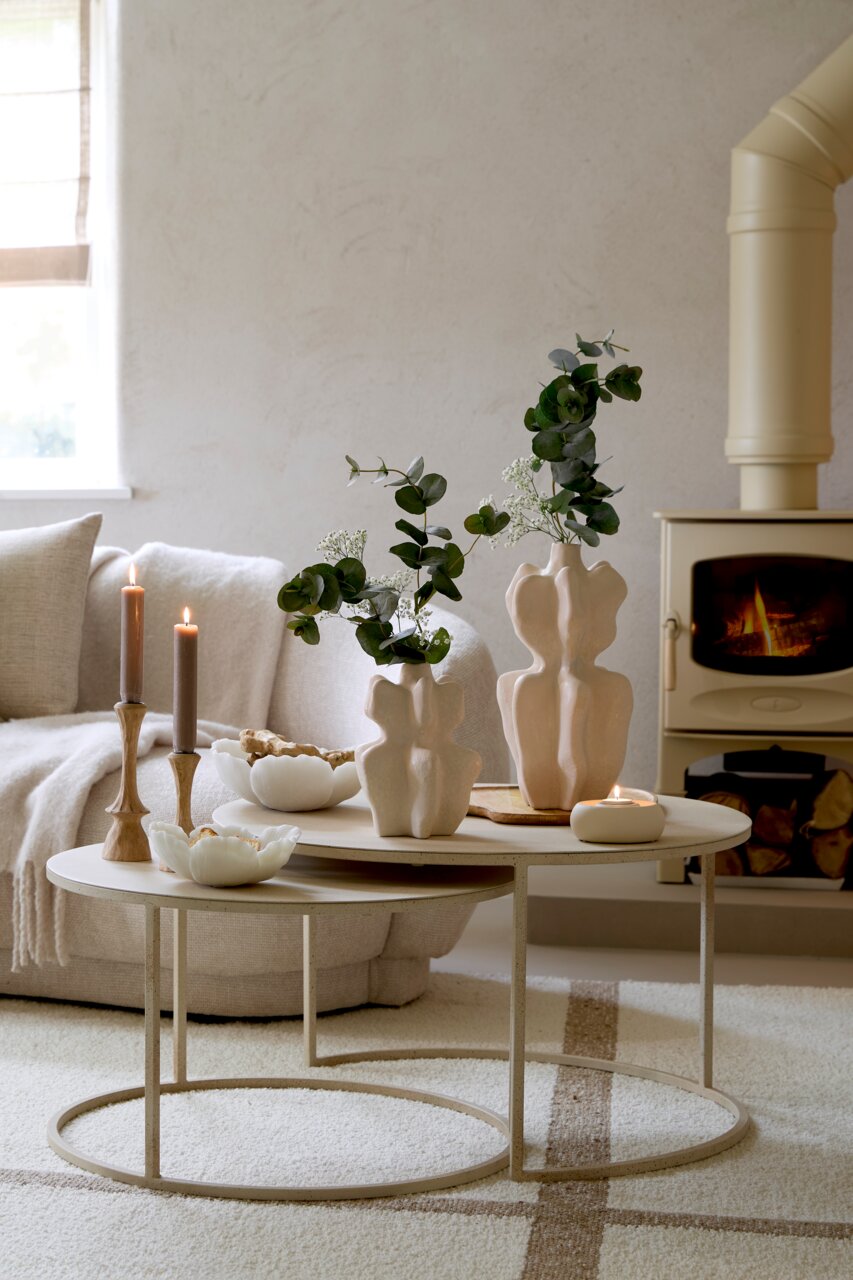 Zwei elegante Kerzenständer aus Holz mit brennenden Kerzen auf einem modernen, runden Couchtisch in einem gemütlichen Wohnzimmer. Perspektive von vorne.