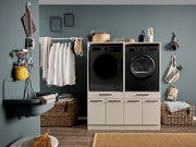 Modern eingerichteter Hauswirtschaftsraum mit schwarzer Waschmaschine und schwarzem Trockner in weißen Unterschränken, Wäsche an einer Stange, Körben und Badewanne; Frontansicht auf die Geräte.