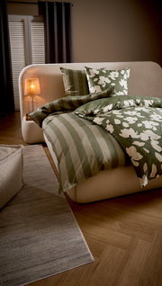 Grün-weißes Bettwäsche-Set mit Streifen- und Blumenmuster auf cremefarbenem Polsterbett, seitliche Perspektive im Schlafzimmer mit Nachttischlampe und Teppich