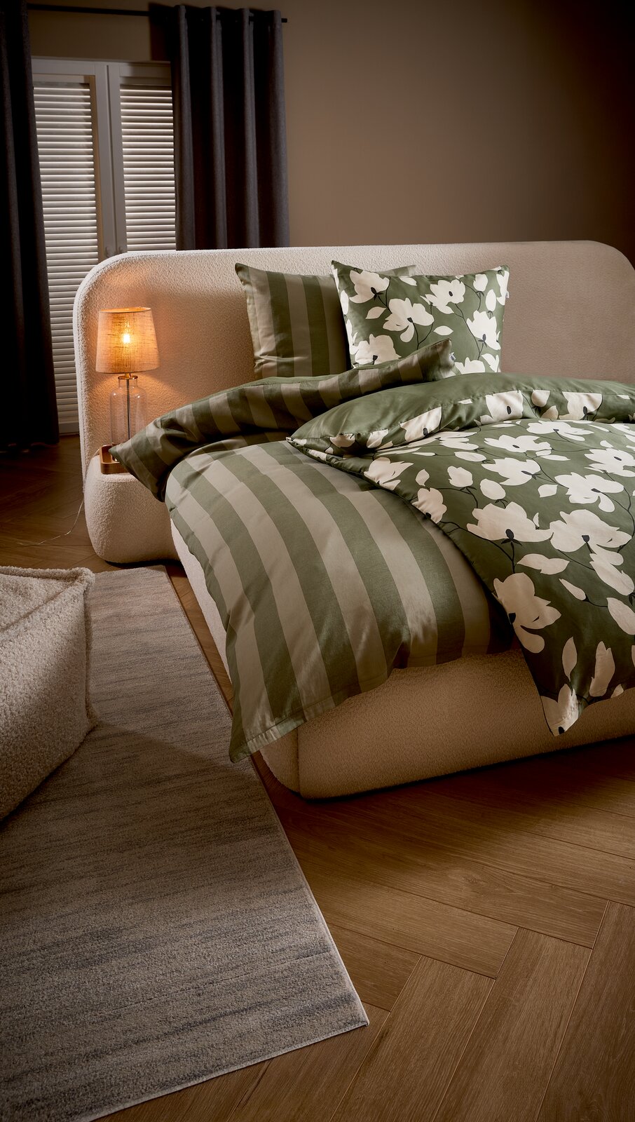 Grün-weißes Bettwäsche-Set mit Streifen- und Blumenmuster auf cremefarbenem Polsterbett, seitliche Perspektive im Schlafzimmer mit Nachttischlampe und Teppich