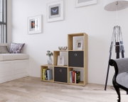 Modernes, stufenförmiges Regal in Eiche-Optik mit offenen Fächern, zwei schwarzen Stoffboxen und Büchern, freistehend an einer weißen Wand im Wohnzimmer; perspektivische Frontansicht leicht von links.