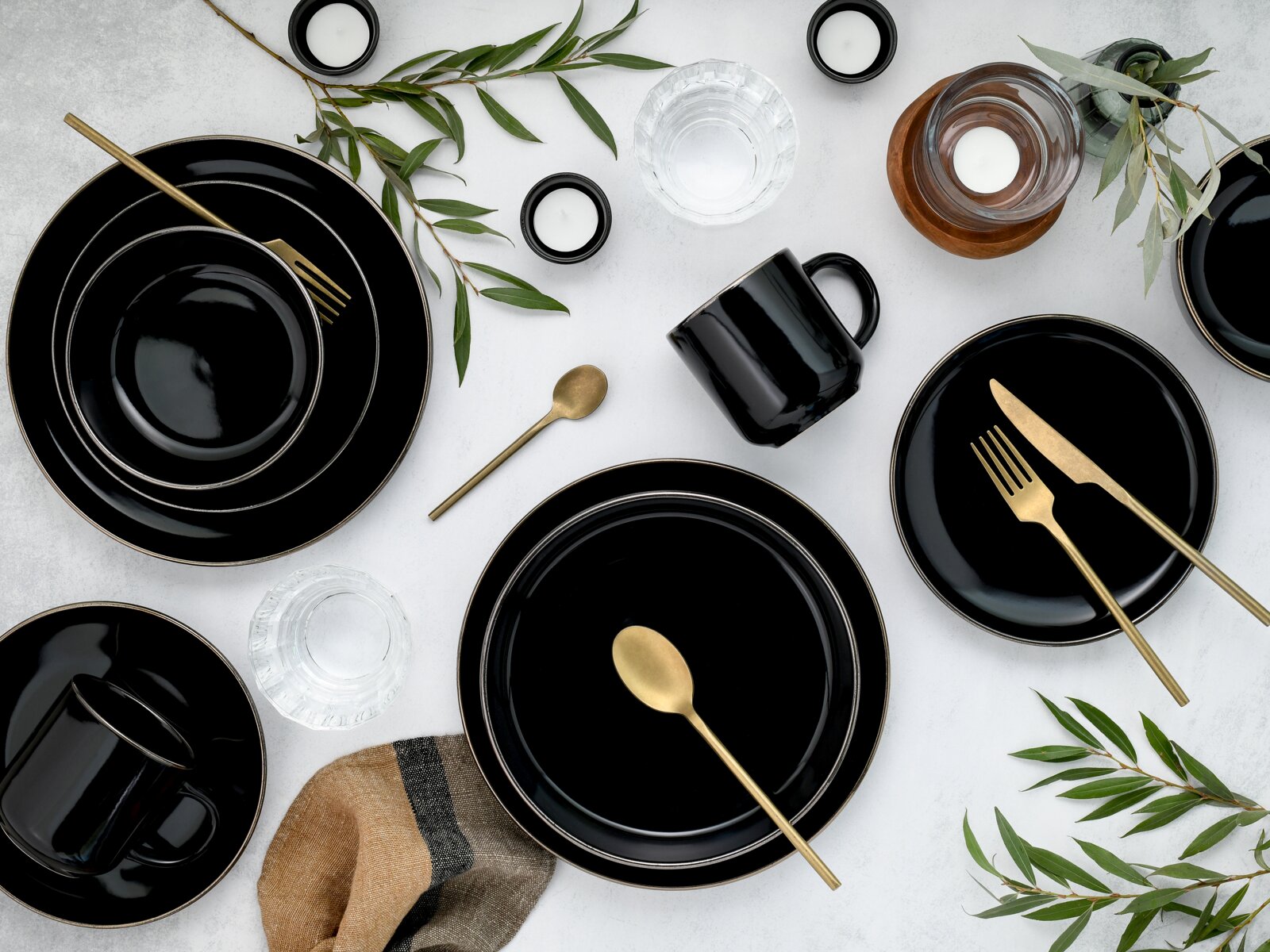 Schwarzes Geschirr-Set mit goldfarbenem Besteck und schwarzen Tassen, dekoriert mit grünen Zweigen und Kerzen auf hellem Tisch – Draufsicht (Flatlay).