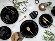 Schwarzes Geschirr-Set mit goldfarbenem Besteck und schwarzen Tassen, dekoriert mit grünen Zweigen und Kerzen auf hellem Tisch – Draufsicht (Flatlay).