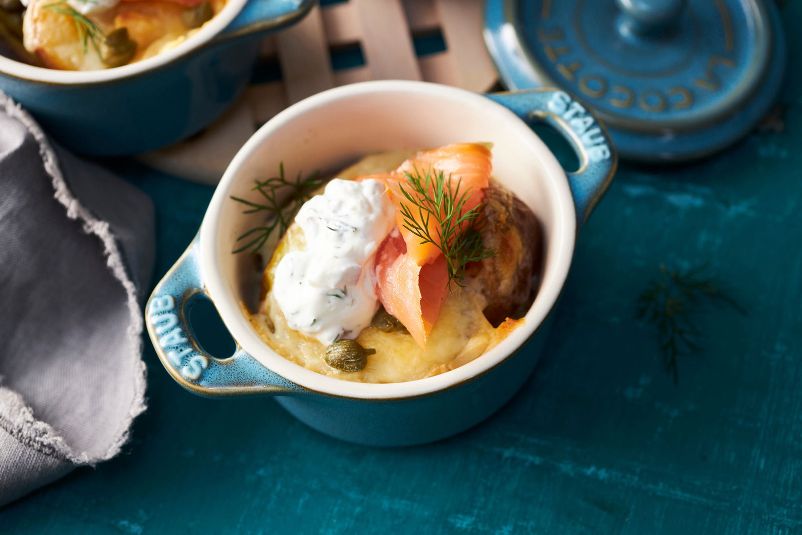 Staub Cocotte Set 4-tlg. Blick von oben auf eine türkisfarbene Mini-Cocotte mit Lachs, Dill und Sahne auf einem blauen Untergrund.