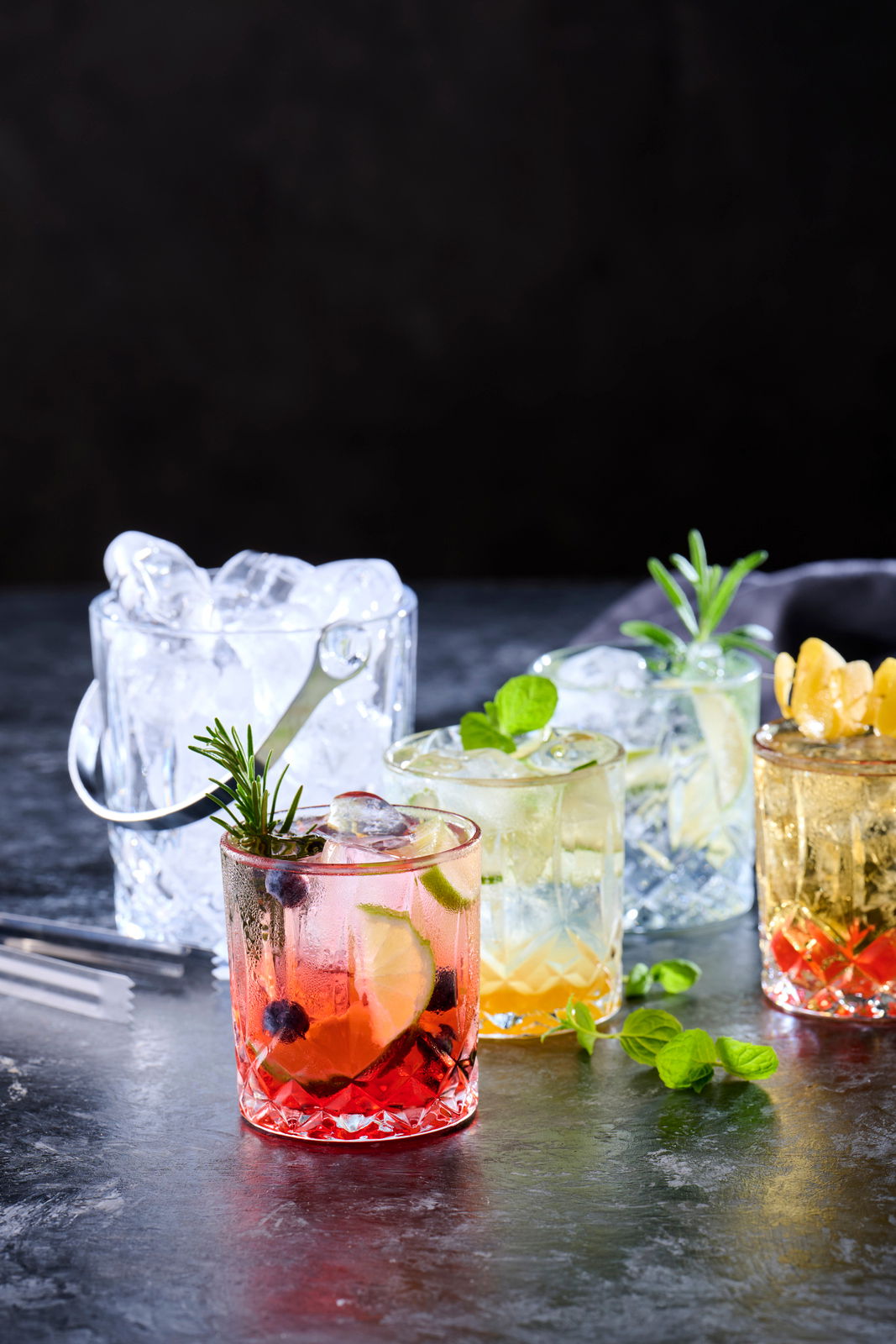 Eiskübel mit Eiswürfeln und vier Gläser mit verschiedenen Getränken, von vorne fotografiert.