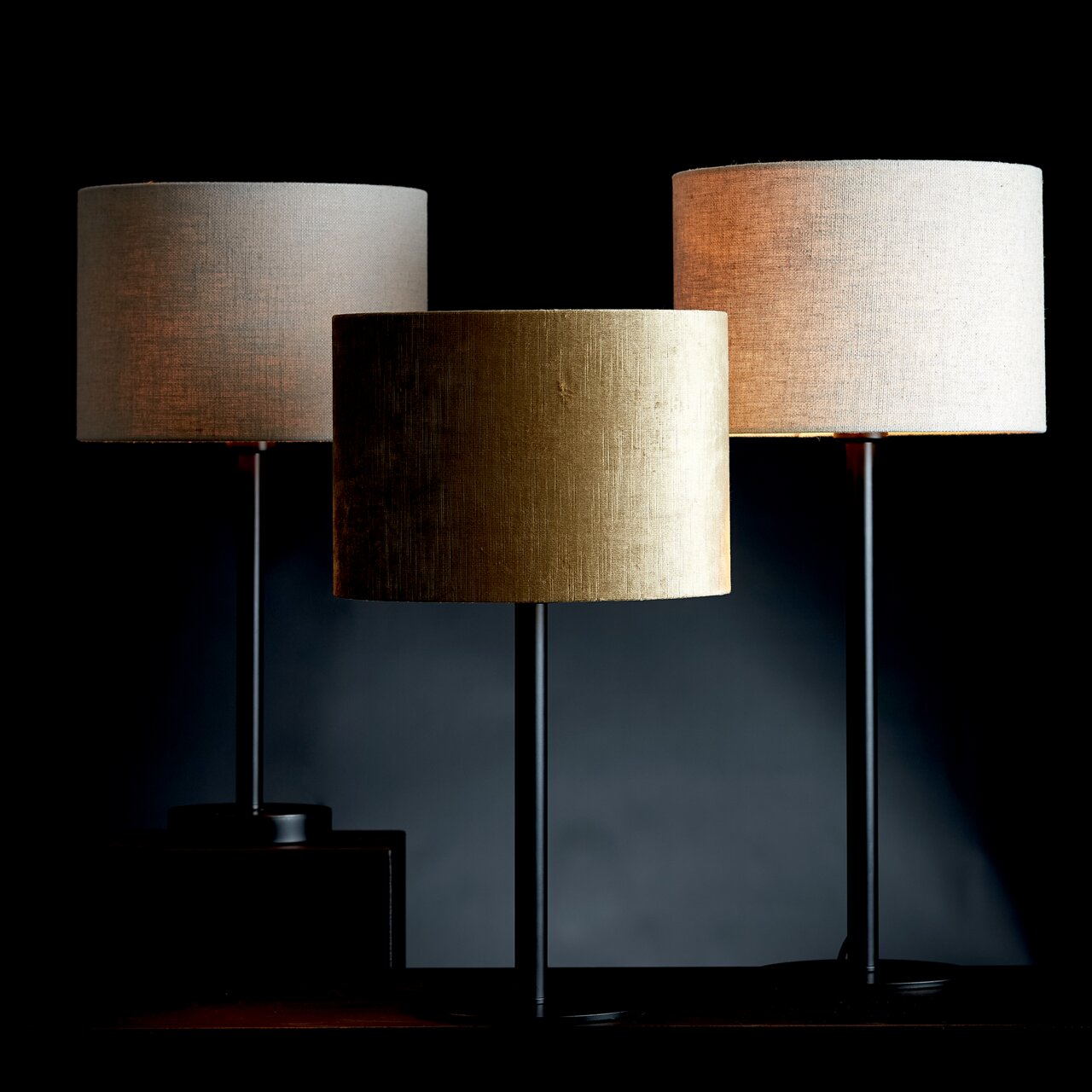 Light&Living Lampenschirm GEMSTONE Drei moderne Lampenschirme in Zylinderform, einer in Gold und zwei in Beige, auf schwarzen Ständern, frontal betrachtet.