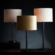 Drei moderne Lampenschirme in Zylinderform, einer in Gold und zwei in Beige, auf schwarzen Ständern, frontal betrachtet.