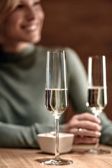 Nahaufnahme eines eleganten Sektglases auf einem Tisch, gefüllt mit Sekt, im Hintergrund eine Person in unscharfer Darstellung.