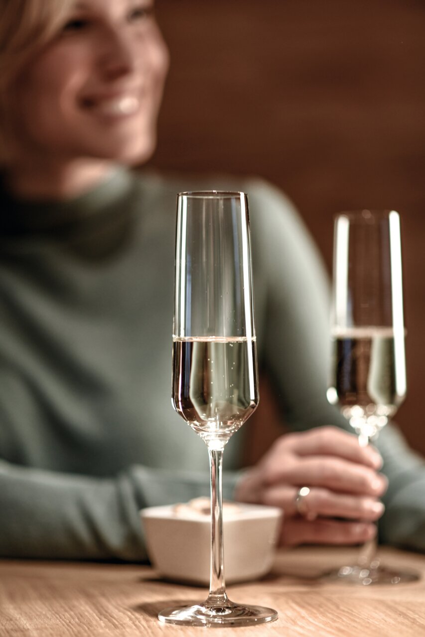 Close-up van een elegant champagneglas op een tafel, gevuld met champagne, met op de achtergrond een persoon die onscherp is weergegeven.