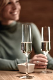 Close-up van een elegant champagneglas op een tafel, gevuld met champagne, met op de achtergrond een persoon die onscherp is weergegeven.
