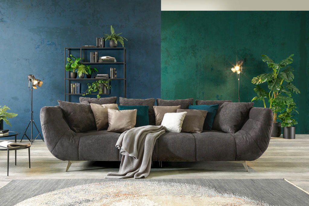 Hom´in Big Sofa FELLINI II. Großes, modernes Sofa in dunklem Stoff mit mehreren Kissen, frontal fotografiert, umgeben von stilvoller Dekoration und Pflanzen.