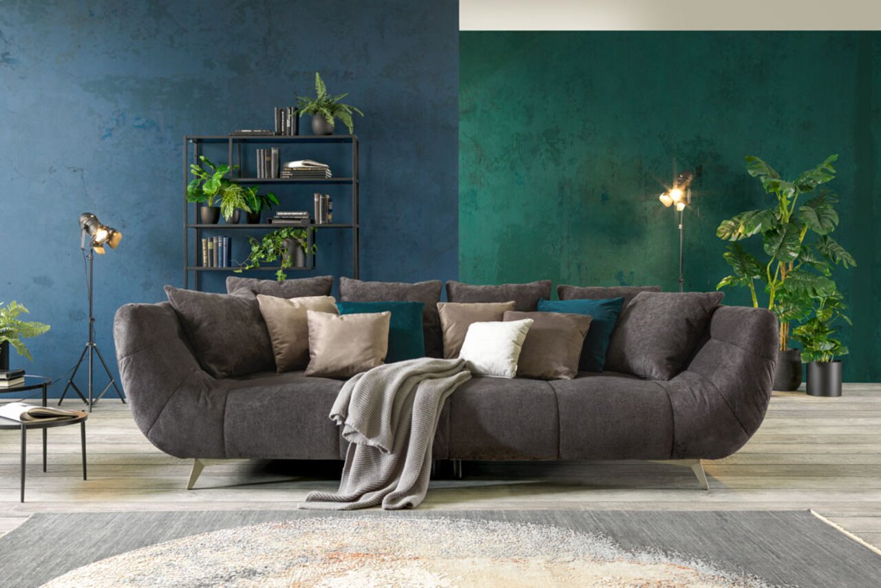Hom´in Big Sofa FELLINI II. Großes, modernes Sofa in dunklem Stoff mit mehreren Kissen, frontal fotografiert, umgeben von stilvoller Dekoration und Pflanzen.