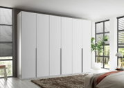 Großer weißer Kleiderschrank mit sechs grifflosen Drehtüren und drei langen schwarzen Griffleisten, aus leicht seitlicher Frontalperspektive in einem hellen modernen Schlafzimmer gezeigt.