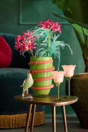 Grüne Vase mit rosa Spiraldekor, gefüllt mit pinken und grünen Kunstblumen, auf einem Holztisch mit Glasvogel und zwei blumenförmigen Gläsern, seitliche Perspektive.