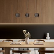 Drei minimalistische Pendelleuchten mit rauchgrauen Glaszylindern hängen in einer Reihe vor Holzpaneelen über einem Esstisch, Frontansicht auf Augenhöhe.