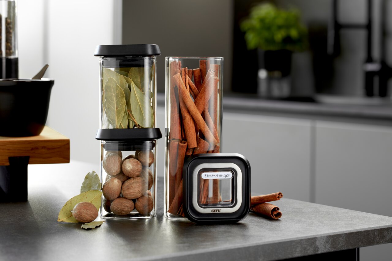 Vorratsdosen-Set PANTRY mit zwei stapelbaren Glasbehältern, gefüllt mit Lorbeerblättern und Muskatnüssen, sowie einem hohen Glasbehälter mit Zimtstangen, auf einer Küchenarbeitsplatte aus der Seitenperspektive.