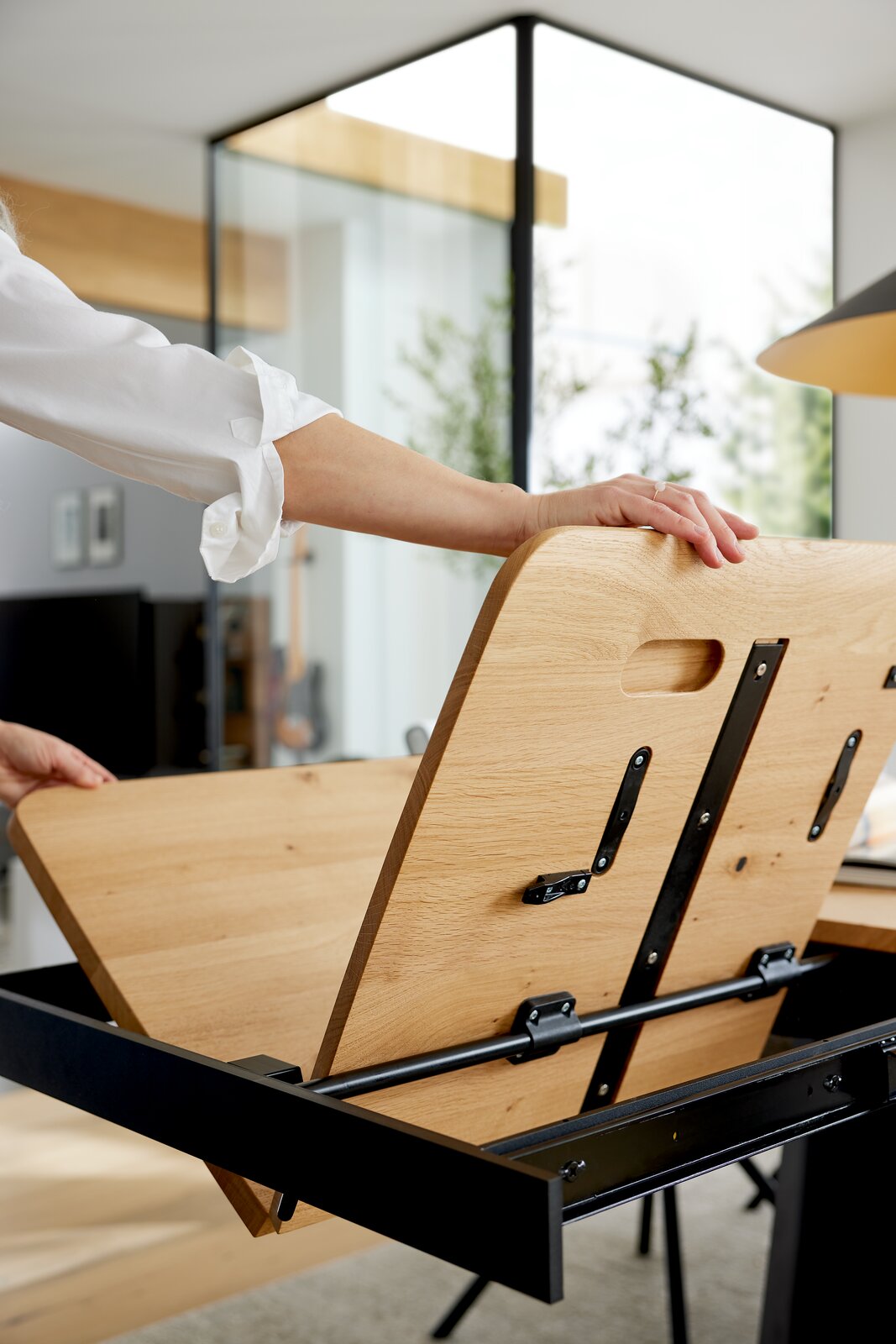 Person klappt eine Holz-Tischplatte mit schwarzem Metallgestell hoch, um den Stauraum darunter zu öffnen; Perspektive: seitliche Nahaufnahme auf die angehobene Tischplatte.