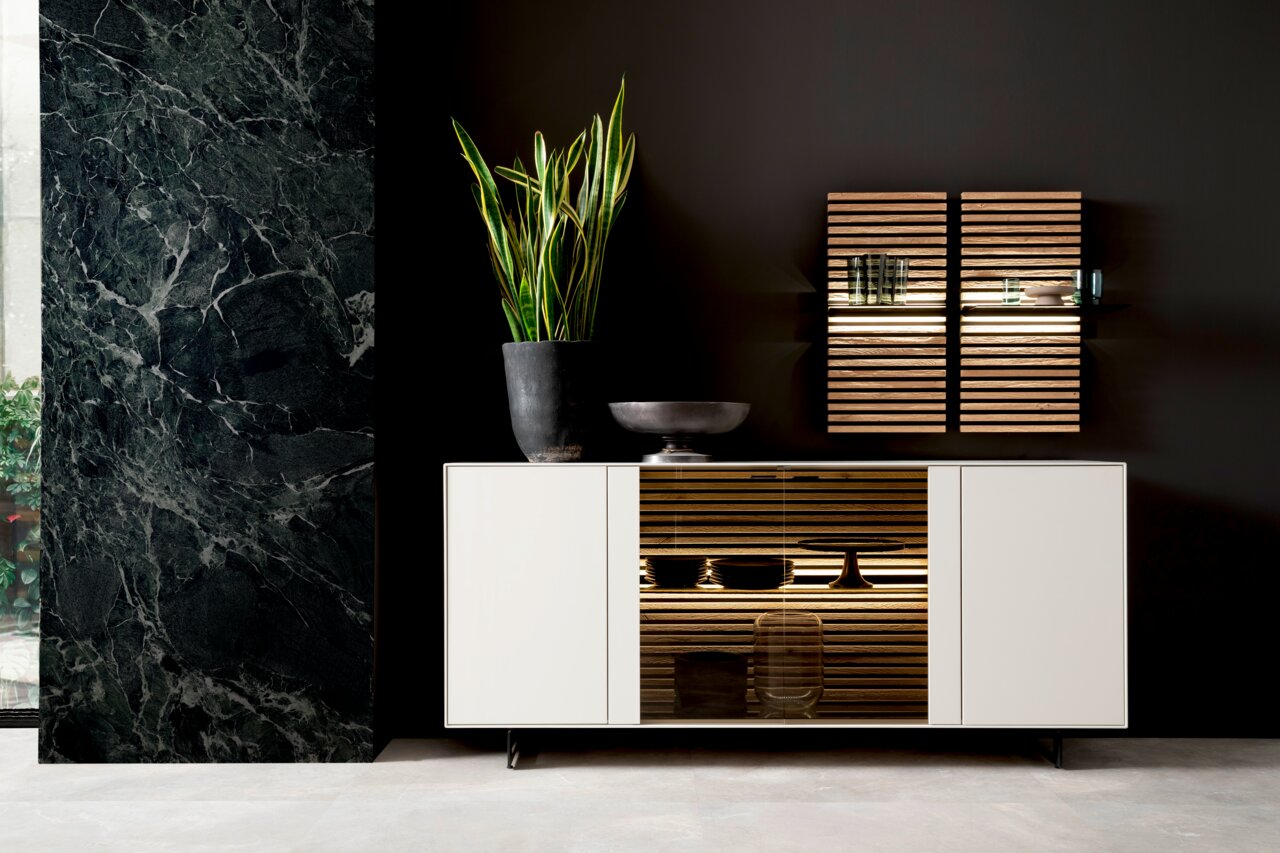 Modernes Sideboard in Weiß mit Glaselementen, dekoriert mit einer Pflanze und einer Schale, vor einer dunklen Wand. Frontalansicht.