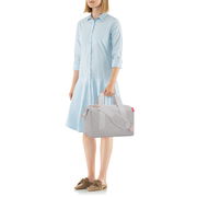 Frau in hellblauem Kleid hält eine graue Allrounder-Tasche in der Hand, seitliche Perspektive